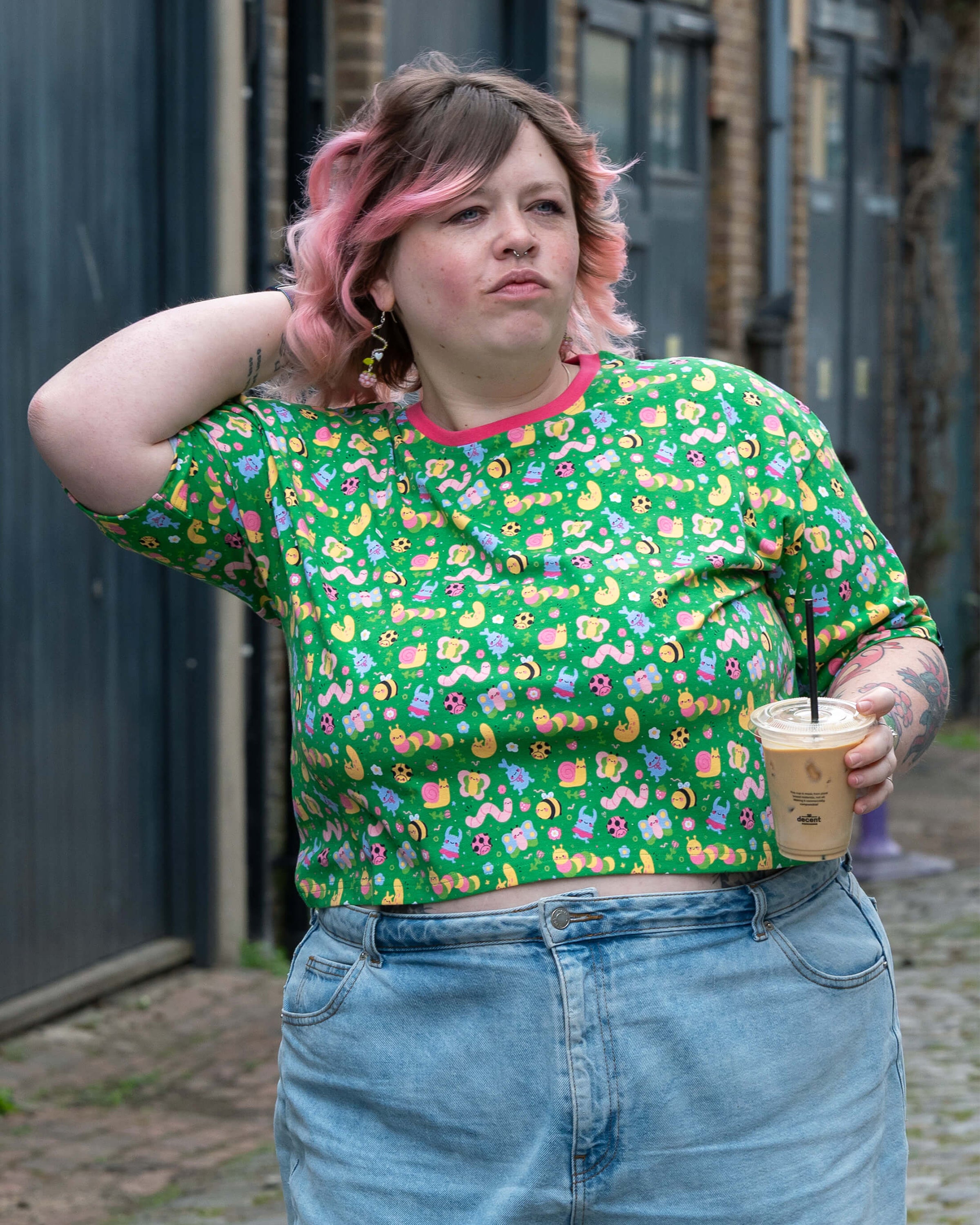 Sam poses with one hand in their hair while holding an iced coffee, wearing the Rebel Romance x Dipstick Store Bugs Cropped Boxy Tee. Styled with light blue high-waisted jeans, the tee’s colourful bug print and pink neckline pop against the brick street background. Rebel Romance UK.