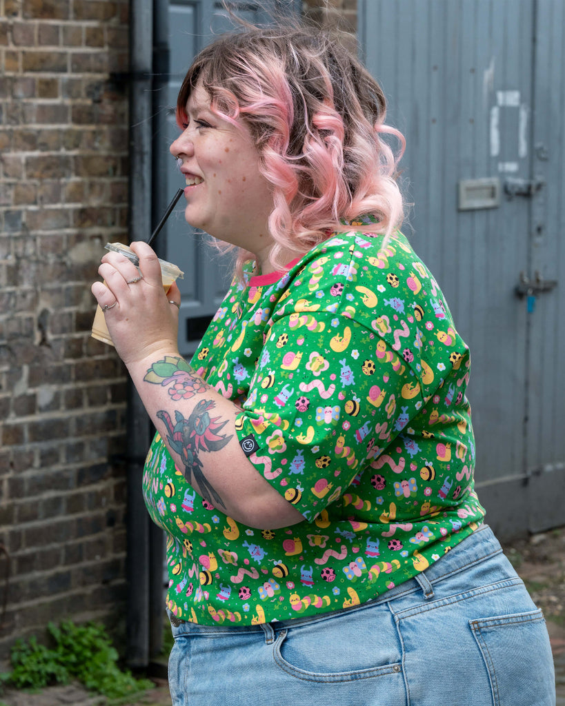 Sam smiles with a drink in hand while wearing the Rebel Romance x Dipstick Store Bugs Cropped Boxy Tee. The green cropped top is covered in cheerful pastel bugs, flowers, and moons, paired with light denim. A playful look from UK ethical brand Rebel Romance.