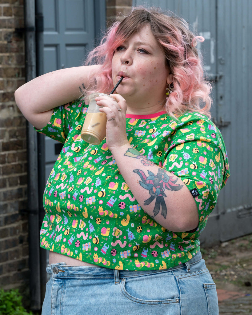 Sam drinks iced coffee while wearing the Rebel Romance x Dipstick Store Bugs Cropped Boxy Tee. The cropped green top with pink collar features a whimsical print of pastel bugs. Styled with high-waisted light blue denim jeans. Inclusive slow fashion by Rebel Romance.