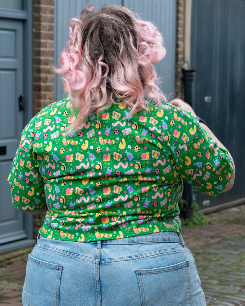 Rear view of Sam, a plus-size model with pastel pink curls, wearing the Rebel Romance x Dipstick Store Bugs Cropped Boxy Tee. The green tee features a fun bug print with snails, worms, moons, and more. Styled with high-waisted light blue denim. Slow fashion by UK brand Rebel Romance.