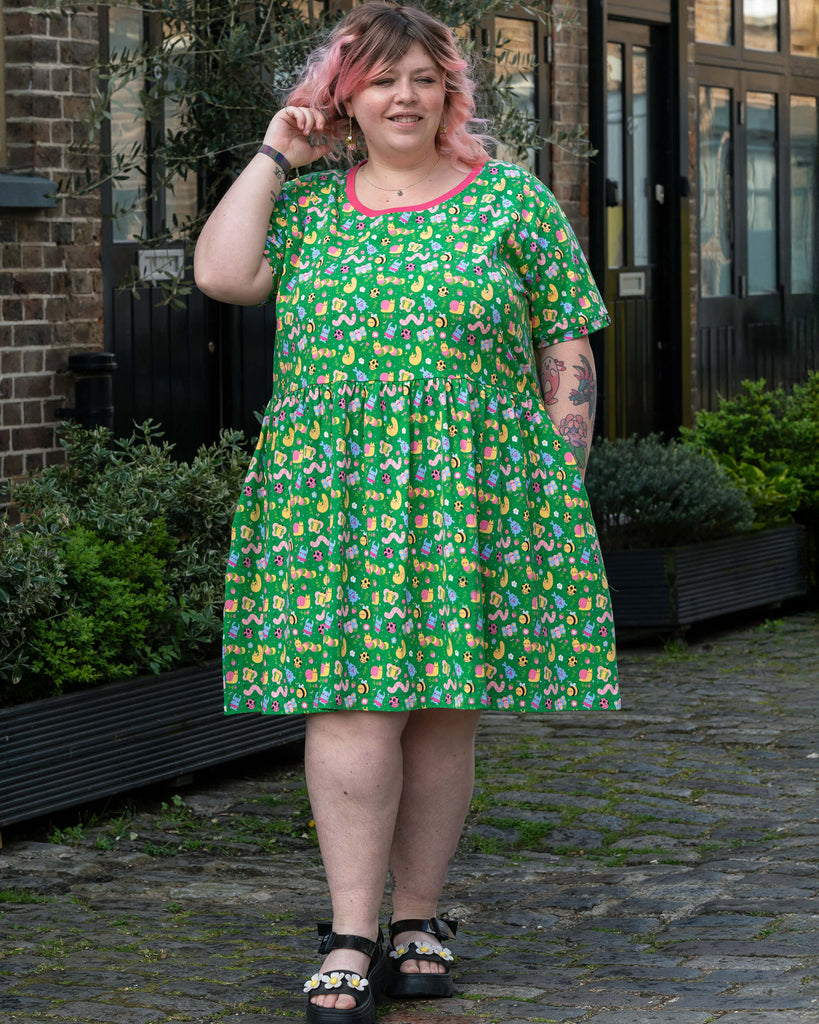 Model with pink and blonde hair holds one hand behind her head and lifts the side of her green bug print smock dress to show its flared shape. The short-sleeved dress features cheerful illustrations of bugs and snails in pastel colours.