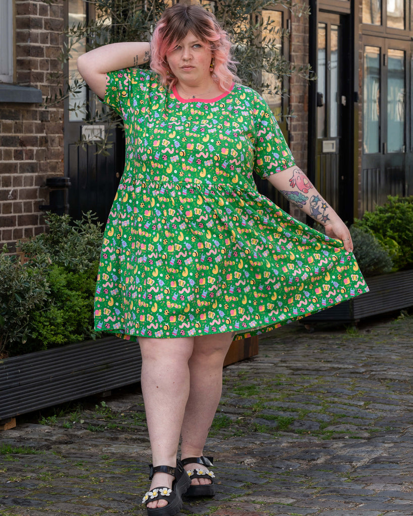 Model with pink ombre curly hair poses on a cobbled street in front of brick buildings, wearing a green stretch smock dress with pink trim and a bright bug-themed print. She accessorises with floral platform sandals and dangly earrings.