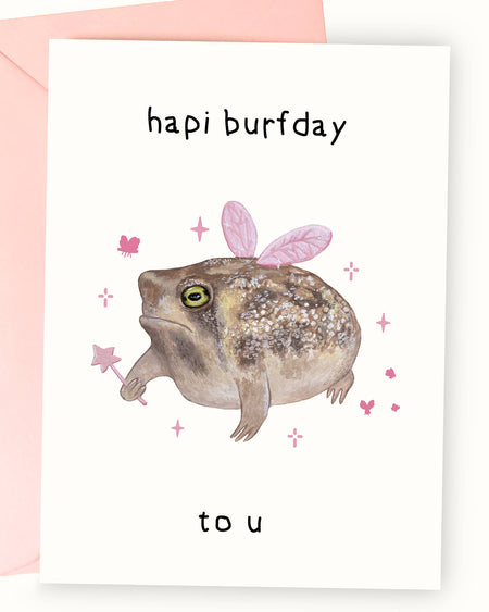 A white greeting card featuring a detailed illustration of a grumpy-looking frog by Nimbob. The frog has textured brown skin and wears soft pink fairy wings on its back. It holds a tiny pink star wand, surrounded by sparkles, butterflies, and a whimsical magical aura. The text above reads ‘hapi burfday’ and below ‘to u’ in a playful, slightly wonky handwritten font. A fun and quirky birthday card perfect for frog lovers and those with a grumpy sense of humour.