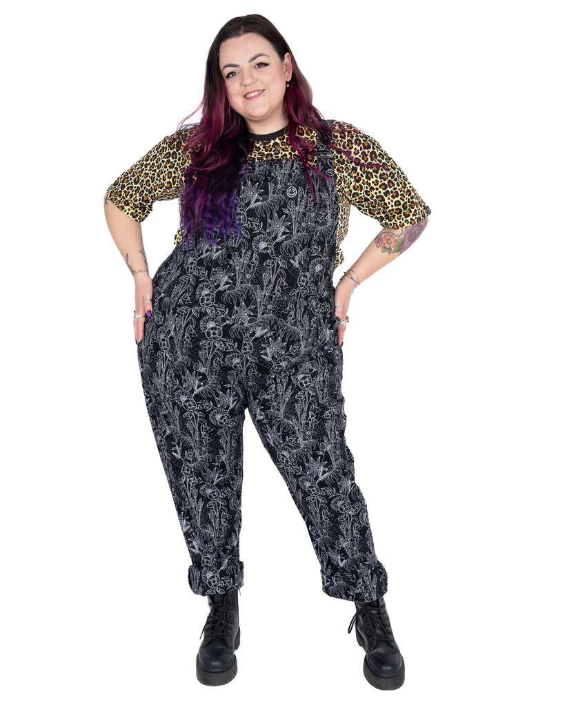 Luisa wears the Rebel Romance Black Floral Cobweb Stretch Dungarees with a confident pose. These comfy, alternative dungarees are decorated with white florals and cobwebs on black stretch fabric for a witchy twist.