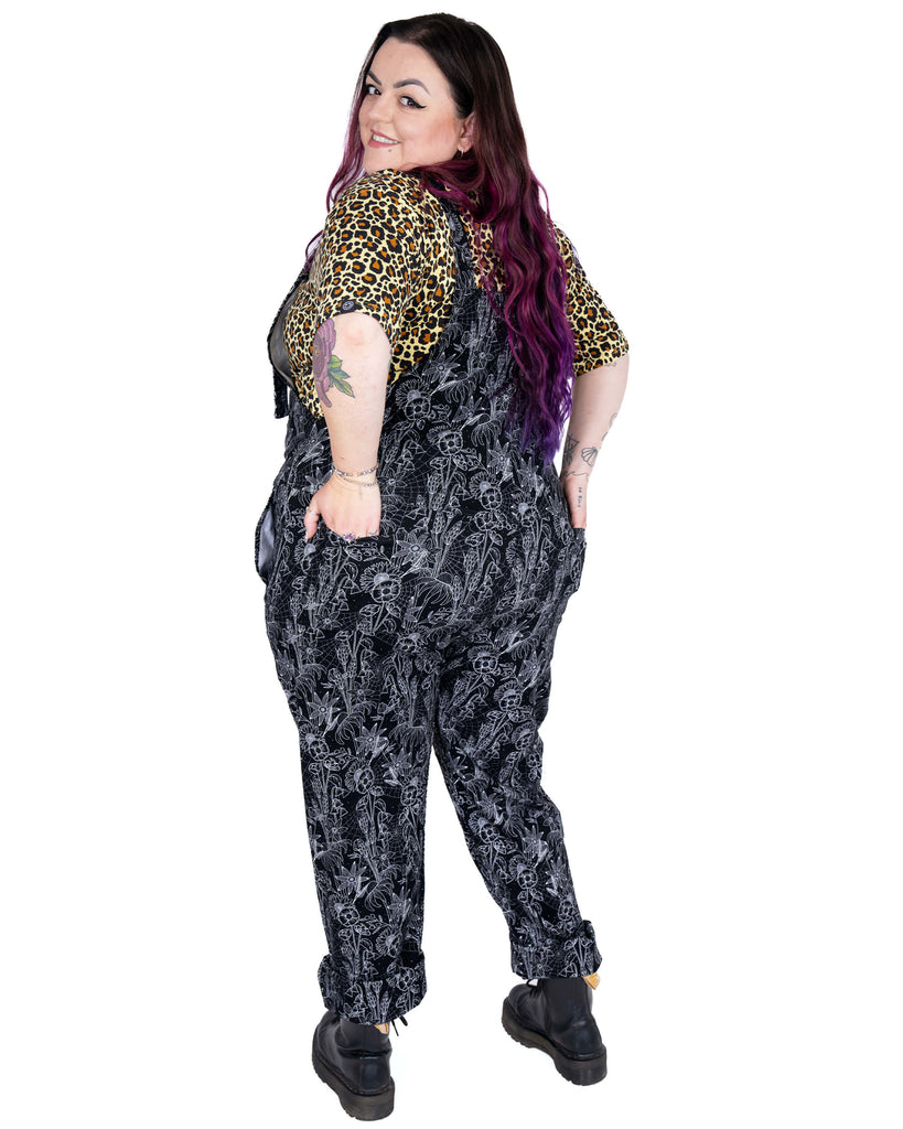 Back view of the Rebel Romance Black Floral Cobweb Stretch Dungarees worn by Luisa. The white botanical and cobweb pattern adds gothic flair to this comfortable and stylish alternative wardrobe staple.