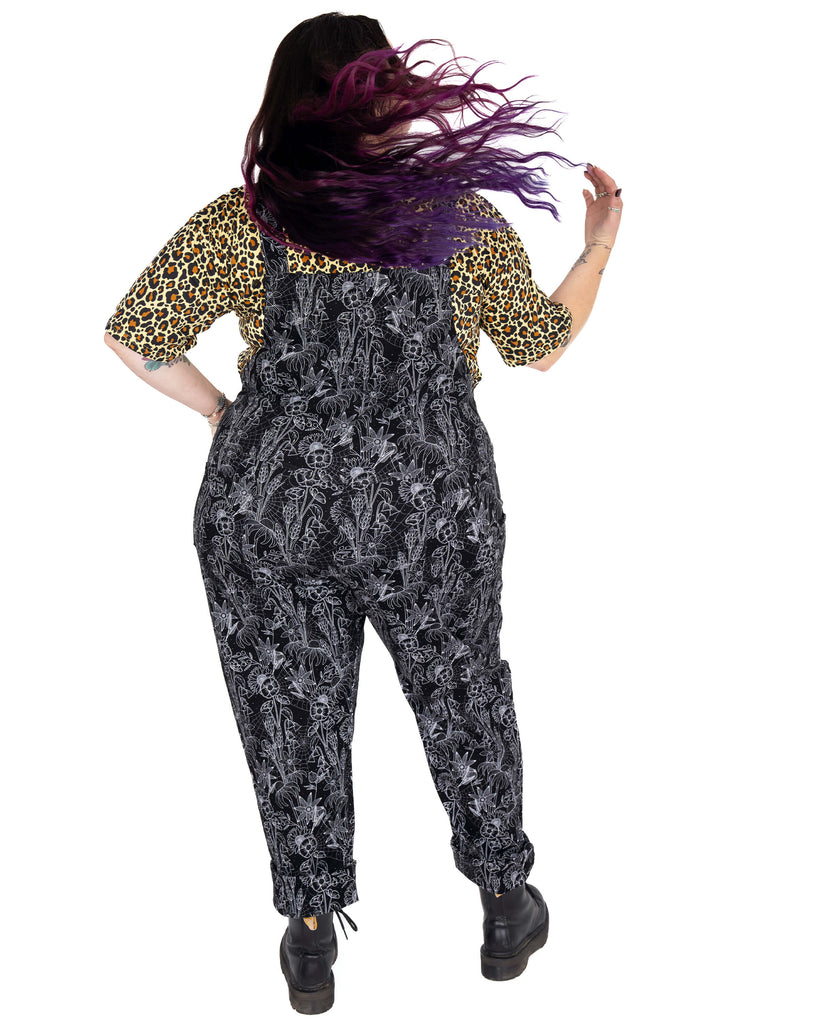 Luisa showcases the back of the Rebel Romance Black Floral Cobweb Stretch Dungarees with flowing purple hair. The all-over white cobweb and floral print pops against the black fabric, perfect for witchy vibes.