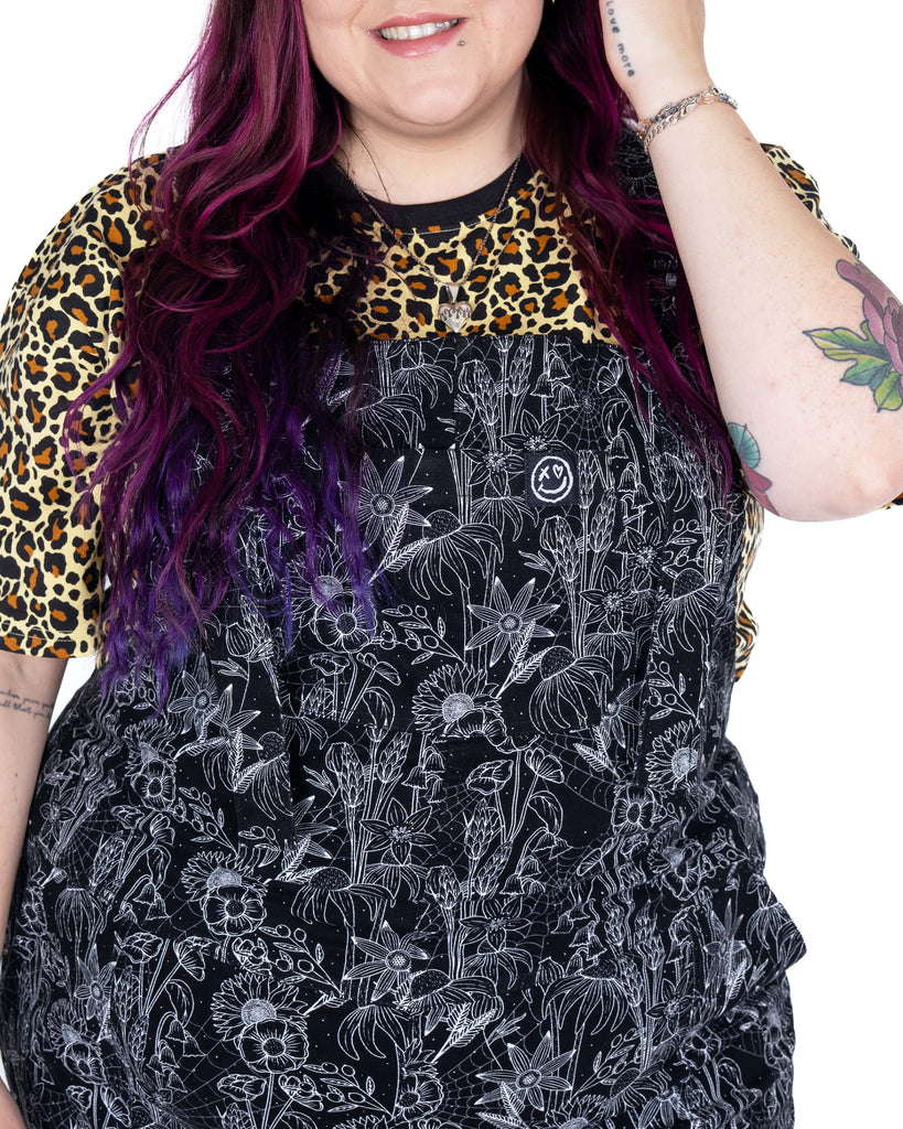 Close-up of the chest bib on the Rebel Romance Black Floral Cobweb Stretch Dungarees, modelled by Luisa. The detailed white floral and spiderweb print is eye-catching and perfect for spooky season style.