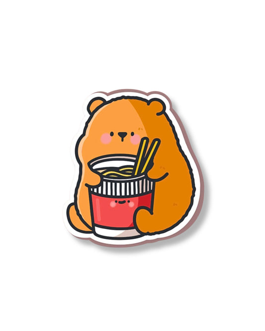 Cartoon bear eating cup noodles sticker with a happy expression, perfect for adding personality to notebooks, seen on a pastel blue surface with matching stationery.