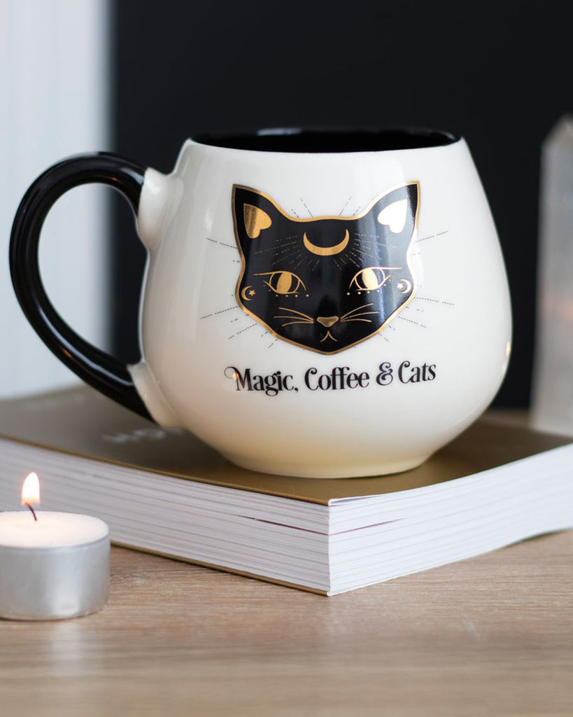 Rounded ceramic mug from Rebel Romance featuring a black and gold mystical cat face design with crescent moon details and the phrase “Magic, Coffee & Cats.” Styled on a book with a tealight candle nearby, perfect for cosy vibes. A witchy, gothic-inspired mug for cat lovers and caffeine fiends from a small UK brand celebrating alternative aesthetics and magical moments.