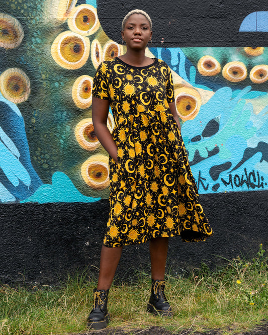 Chish wearing a black and yellow patterned celestial midi smock dress standing in front of a colourful mural.