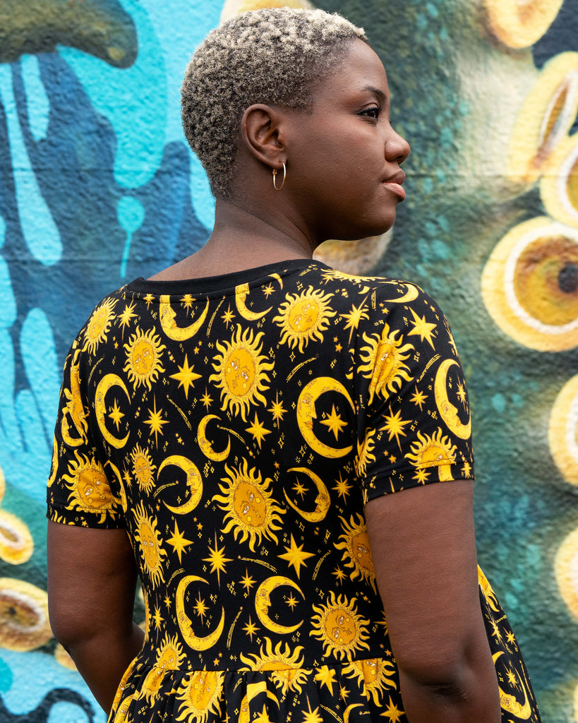 Back view of Chish modelling the Rebel Romance Celestial Midi Dress in stretch cotton. The black smock dress is printed with yellow suns, crescent moons, and stars for a celestial, witchy-inspired alternative fashion look.