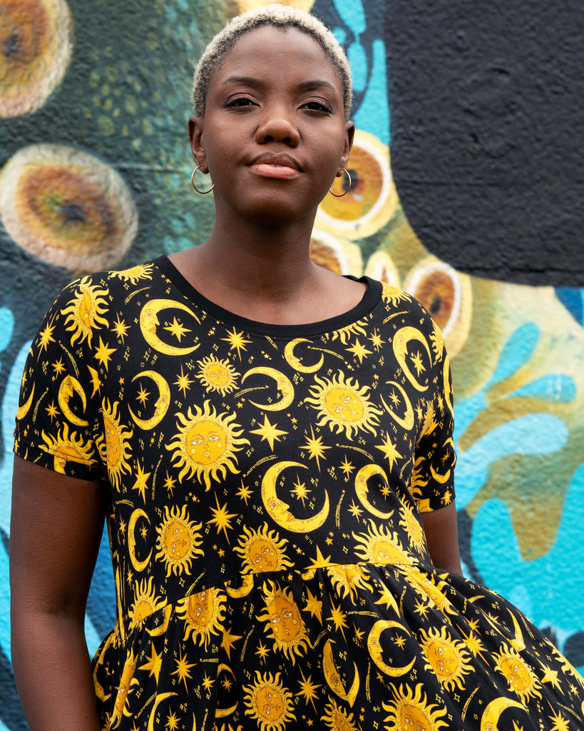 Close-up of Chish modelling the Rebel Romance Celestial Midi Dress in stretch cotton. The celestial print shows golden suns, crescent moons, and stars on a black background, creating a striking witchy and alternative look.