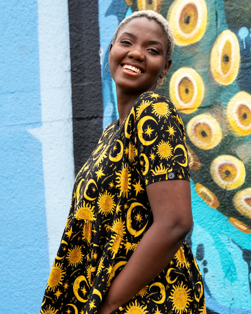 Side close-up of Chish modelling the Rebel Romance Celestial Midi Dress. The yellow celestial print with moons, suns, and stars stands out on black stretch cotton fabric. A comfy and stylish midi dress for alternative and witchy wardrobes.