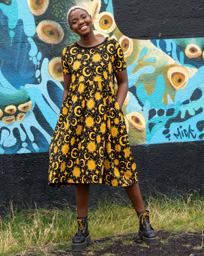 Chish modelling a black and yellow patterned celestial smock midi dress standing in front of a colourful mural.