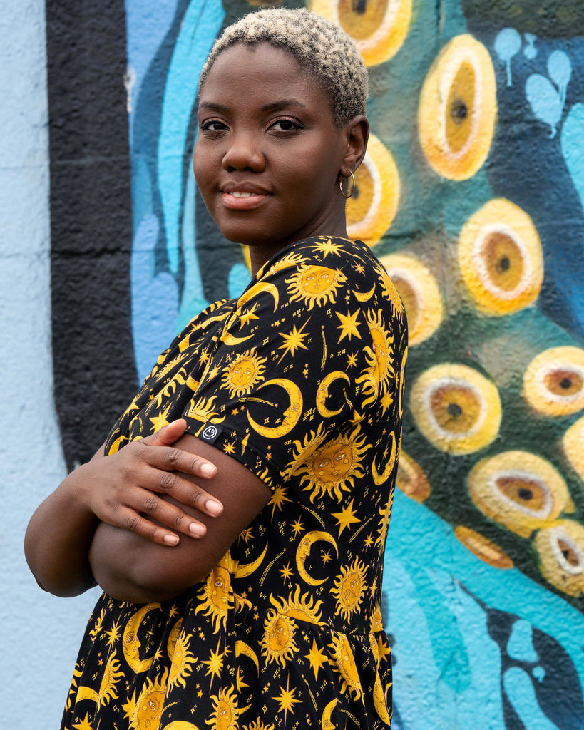 Side close-up of Chish modelling the Rebel Romance Celestial Midi Dress. The yellow celestial print with moons, suns, and stars stands out on black stretch cotton fabric. A comfy and stylish midi dress for alternative and witchy wardrobes.