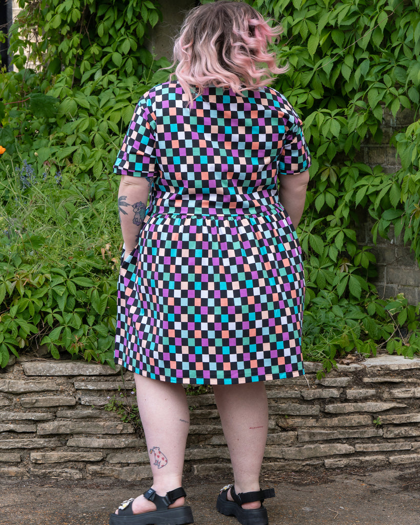 Back view of Sam wearing the Rebel Romance Checkerboard Stretch Smock Dress. This colourful smock dress features a playful pastel checkerboard print and a relaxed fit. Sam is a UK size 22–24 and 5'3, shown here outdoors against a leafy green backdrop. Ethically made by UK brand Rebel Romance, which celebrates alternative fashion and bold style.