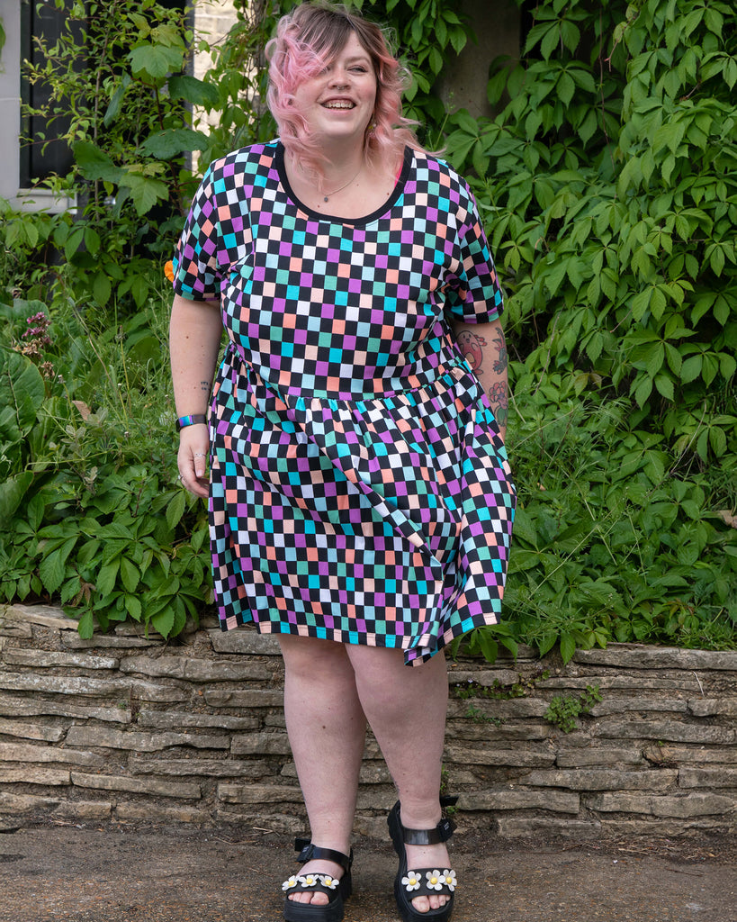 Front view of Sam smiling in the Rebel Romance Checkerboard Stretch Smock Dress. The dress features short sleeves, a black neckline trim, and a bold multicolour checkerboard print. Sam, a UK size 22–24, wears a size 24. Rebel Romance is a small UK brand celebrating slow ethical fashion and inclusive, alternative style.