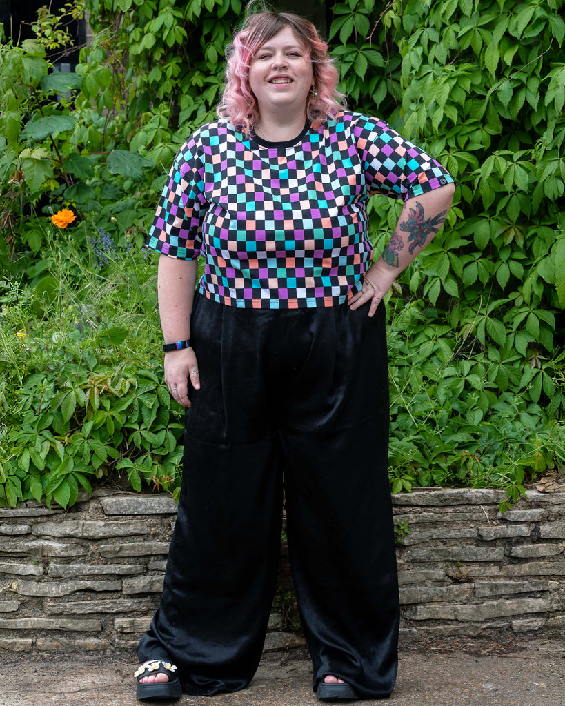 Full-body shot of Sam wearing the Rebel Romance pastel checkerboard tee with black velvet wide-leg trousers and platform sandals. The colourful outfit pops against the green background and garden wall.
