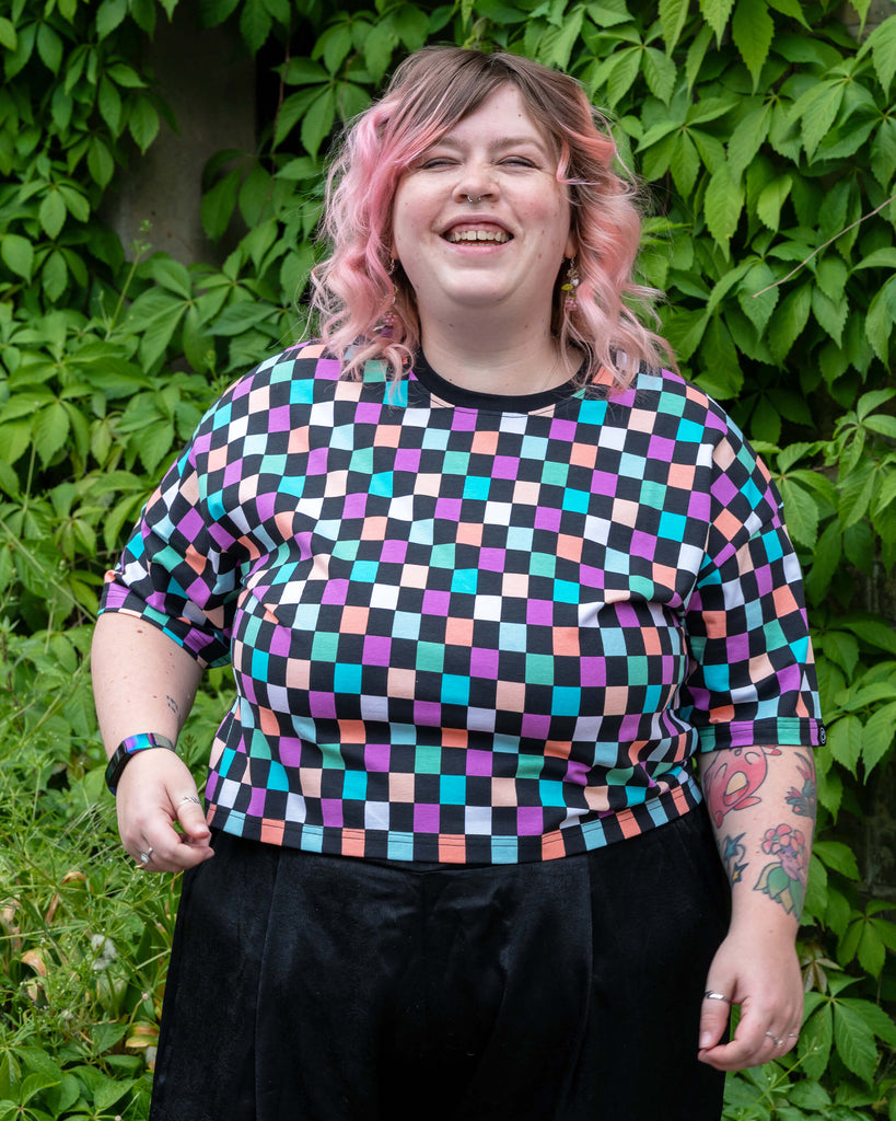 Front-facing image of Sam smiling brightly in the Rebel Romance Checkerboard Boxy Tee, styled with black wide-leg trousers. The colourful check print and lilac neckline stand out beautifully against the green foliage.