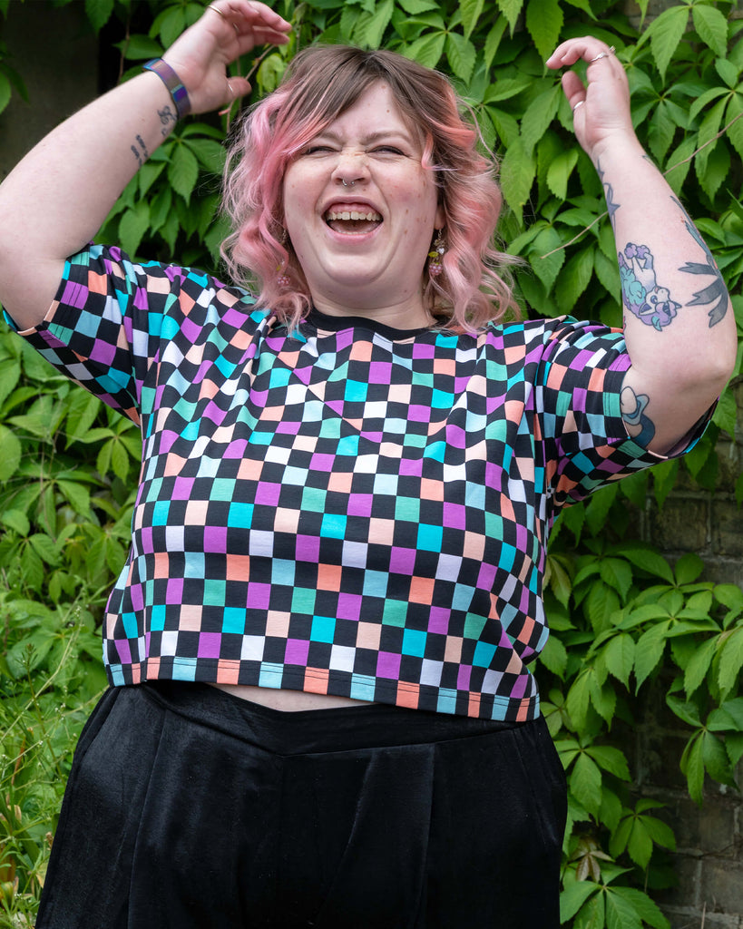 Close-up of Sam mid-laugh in the Rebel Romance Checkerboard Boxy Tee, arms raised with visible tattoos and pastel curls. The cropped fit and bold colourful design channel dopamine dressing energy.