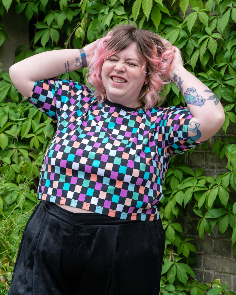 Sam, wearing a pastel checkerboard cropped tee by Rebel Romance, lifts their hands into their pink curly hair and laughs. The oversized boxy fit and black velvet trousers create a fun, relaxed aesthetic against the leafy background.