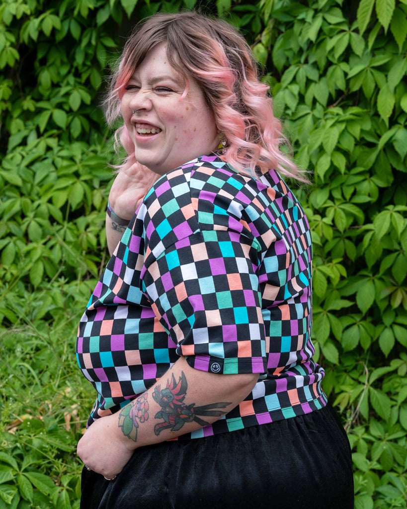 Sam, a UK size 22-24 model, poses side-on with a joyful expression, wearing the Rebel Romance Checkerboard Boxy Tee featuring multicoloured pastel squares. Set against lush green ivy, their pink hair and tattoos add to the bold alternative vibe.