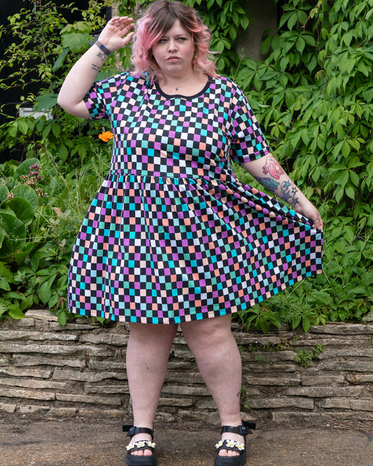 Sam poses confidently in the Rebel Romance Checkerboard Stretch Smock Dress with hands holding out the skirt. The dress features a pastel checkerboard print on black, gathered waist, and relaxed fit. Sam is a UK size 22–24 and 5'3, wearing a 24. Rebel Romance is a UK brand for bold, ethical, alternative fashion lovers.