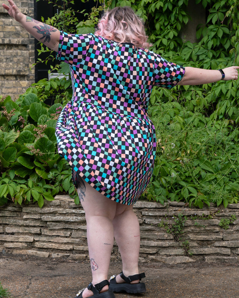 Playful motion shot of Sam spinning in the Rebel Romance Checkerboard Stretch Smock Dress, showcasing the flowy shape and vibrant checkerboard print. Designed for comfort and self-expression, this ethical smock dress is made by Rebel Romance, a UK brand known for alternative and inclusive fashion.
