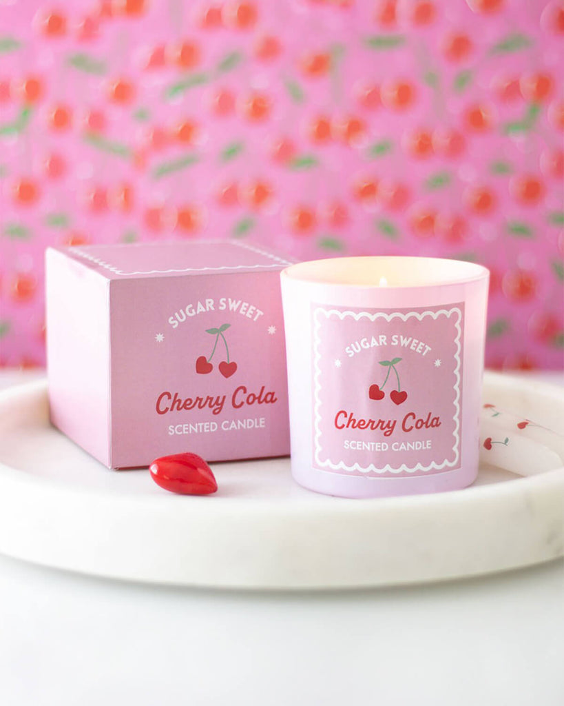 Lifestyle shot of the Sugar Sweet Cherry Cola Candle lit, placed on a white tray with cherry-themed décor in the background.