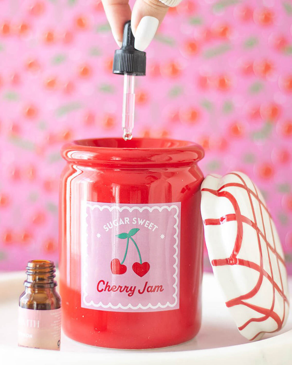  A hand dripping essential oil into the cherry jam jar oil burner with a dropper, against a background with pink and cherry-themed wallpaper.