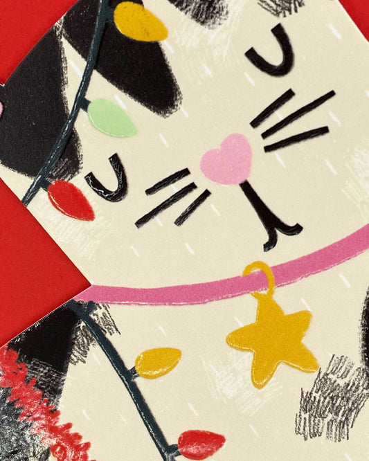 Black and white cat Christmas card featuring a cheeky kitty tangled in red tinsel and multicoloured fairy lights. The die-cut design is finished with embossing and spot UV gloss for a tactile effect, perfect for cat lovers this festive season.