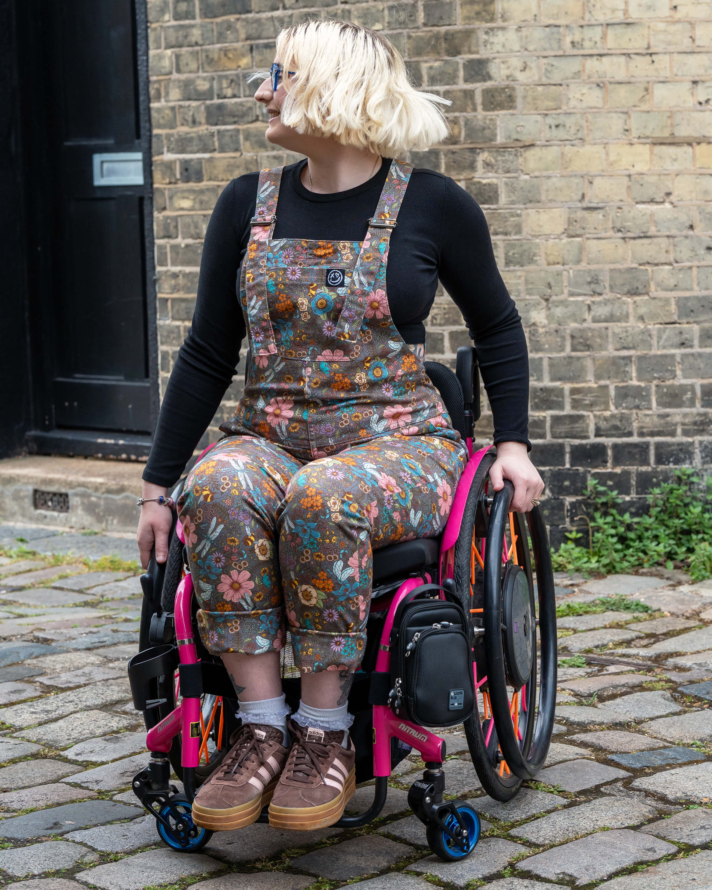 Eliza, a non-binary model and wheelchair user, wears Rebel Romance’s Cottagecore Stretch Twill Dungarees outdoors. Featuring a vintage-style floral woodland print, the dungarees are paired with a black top and brown trainers. A bold and inclusive look from UK indie label Rebel Romance.