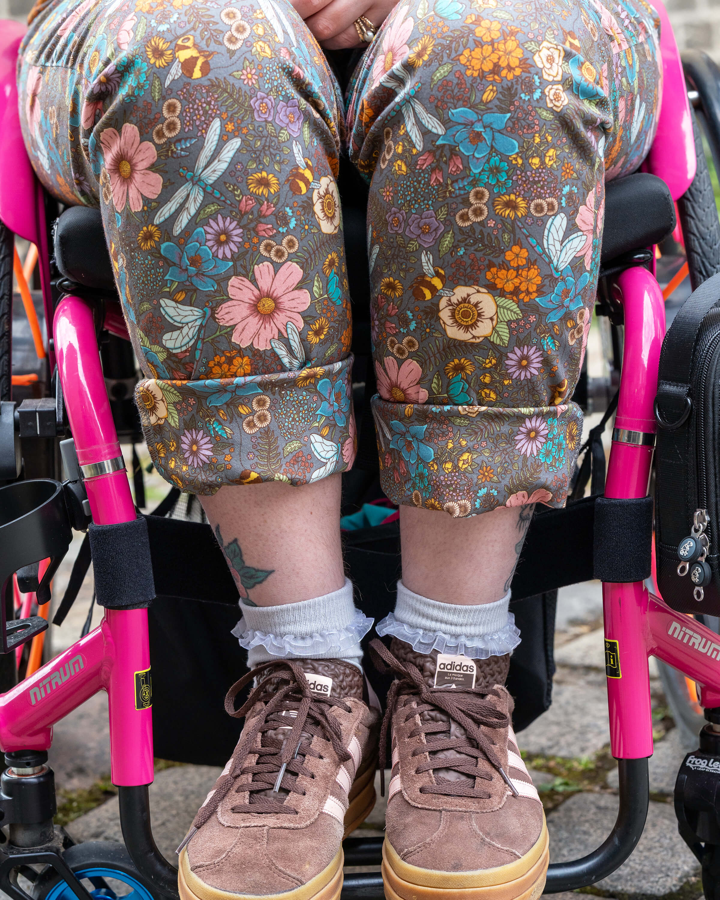 Close-up of the Rebel Romance Cottagecore Stretch Twill Dungarees worn by Eliza, seated in a pink wheelchair. The turned-up legs show a floral woodland print with bees and dragonflies. Styled with frilly socks, brown Adidas trainers, and visible leafy tattoos. A cheerful ethical fashion choice from UK brand Rebel Romance.