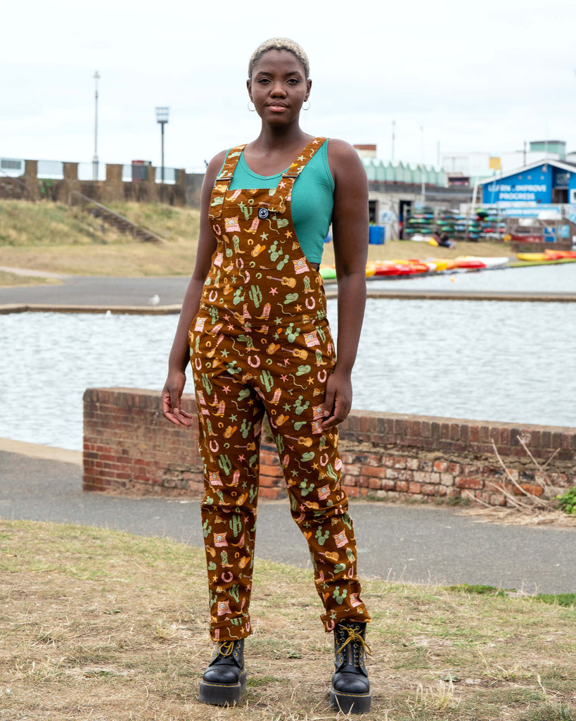 Model by the water in Rebel Romance x Yasmin Cousins Cowboy Stretch Dungarees featuring a western print with cacti, boots, guitars, snakes and sheriff hats, made from stretch twill in a unisex fit.