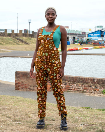 Model by the water in Rebel Romance x Yasmin Cousins Cowboy Stretch Dungarees featuring a western print with cacti, boots, guitars, snakes and sheriff hats, made from stretch twill in a unisex fit.