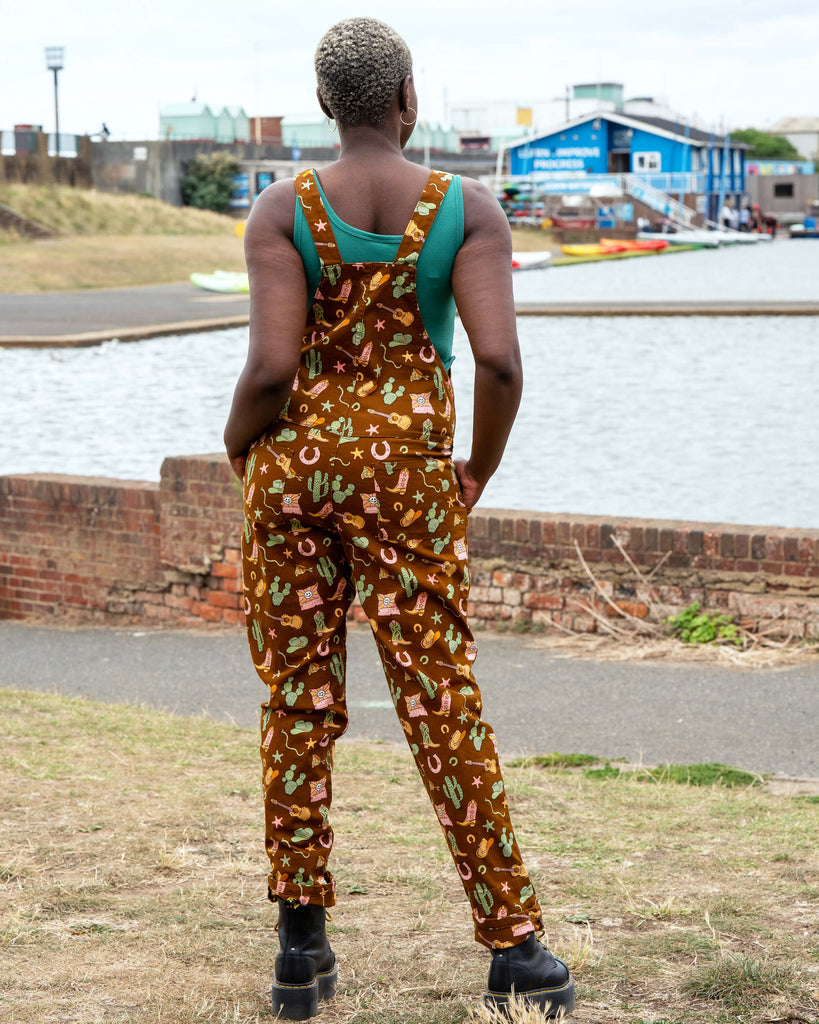Back view of Rebel Romance x Yasmin Cousins Cowboy Stretch Dungarees in brown stretch twill, printed with playful cowboy details including cacti, snakes, boots and guitars.
