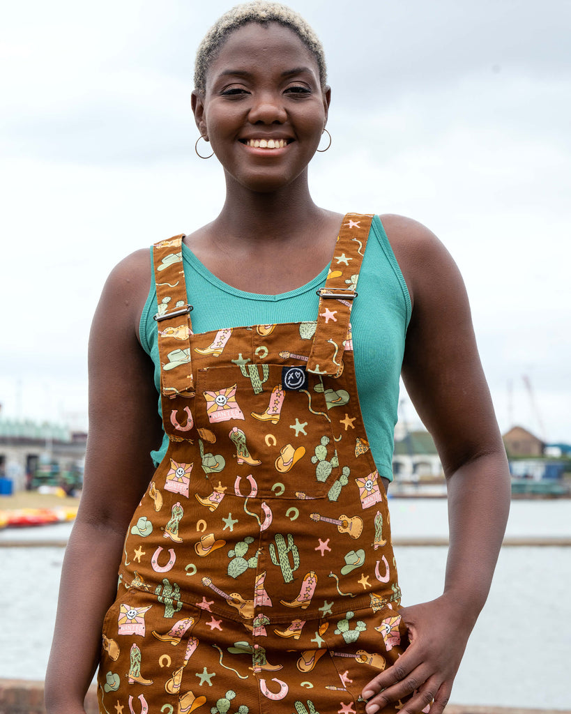 Model smiling in Rebel Romance x Yasmin Cousins Cowboy Dungarees, a quirky unisex fit with colourful western-themed print on comfy stretch twill.