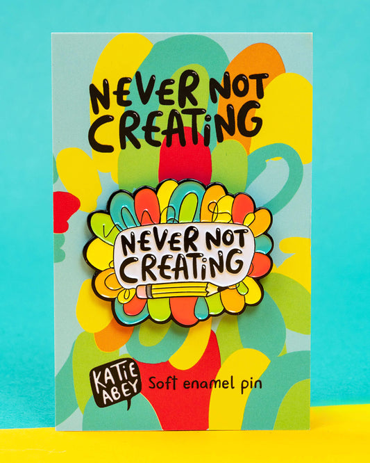 A colourful enamel pin badge by Katie Abey reading “Never Not Creating,” displayed on its backing card. The backing features bright rainbow doodles and bold black text. Sold by Rebel Romance, a UK brand celebrating alternative fashion, gifts and homeware for creative millennials.