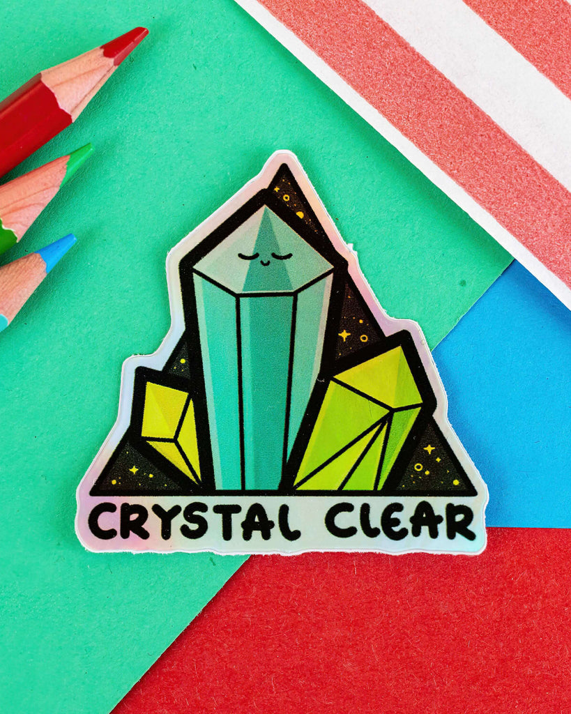 Crystal Clear Holographic Sticker by Rebel Romance, featuring a cute illustrated crystal cluster with a smiling face and text that reads "Crystal Clear." Designed by Innabox, this waterproof vinyl sticker sparkles with a holographic finish, perfect for decorating laptops, water bottles, and notebooks. A vibrant and spiritual sticker for alternative millennials who love crystals.