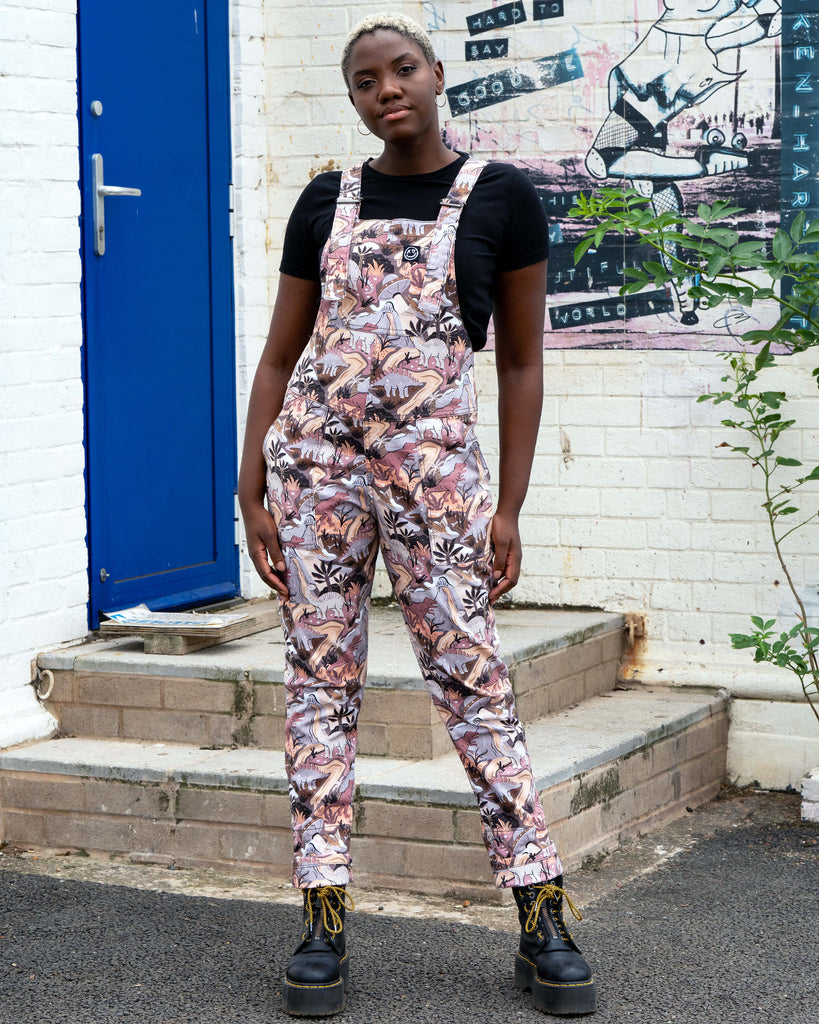 Femme person with short white blonde hair wearing the Rebel Romance dinoverse dinosaur dungarees, a print in grey, warm oranges and pastel red of dinosaurs, rivers, plants and a hidden UFO. She is standing in front of a blue door and brick wall with graffiti.
