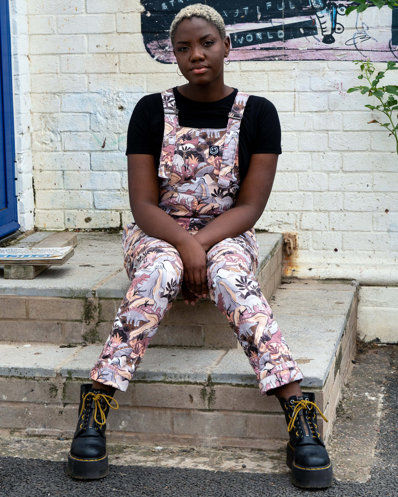 Femme person with short white blonde hair wearing the slow ethical fashion brand Rebel Romance dinoverse dinosaur dungarees, a print in grey, warm oranges and pastel red of dinosaurs, rivers, plants and a hidden UFO. She is sat on some steps in front of a blue door and brick wall with graffiti.