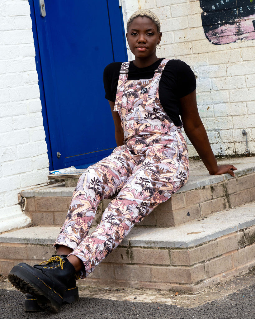 Femme person with short white blonde hair wearing the Rebel Romance dinoverse dinosaur dungarees, a print in grey, warm oranges and pastel red of dinosaurs, rivers, plants and a hidden UFO. She is sat down in front of a blue door and brick wall with graffiti.