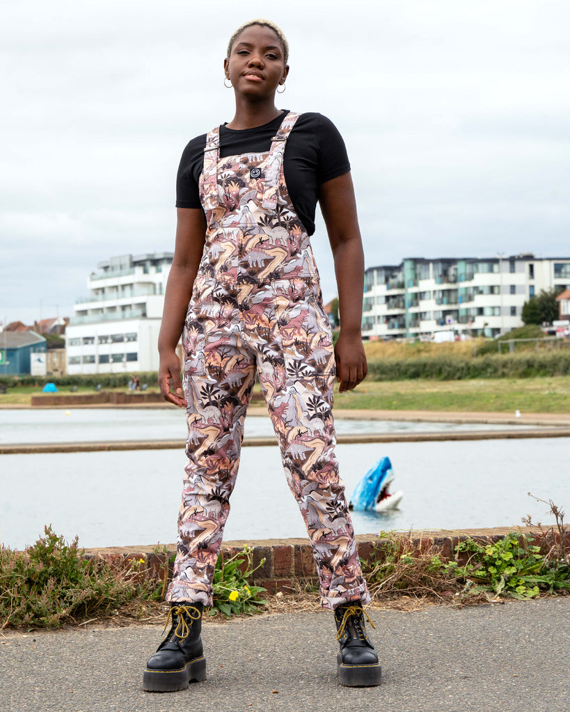 Femme person with short white blonde hair wearing dopamine dressing brand Rebel Romance dinoverse dinosaur dungarees, a print in grey, warm oranges and pastel red of dinosaurs, rivers, plants and a hidden UFO. She is standing by a waterfront with buildings in the background.