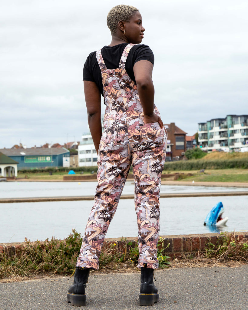 Femme person with short white blonde hair wearing dopamine dressing brand Rebel Romance dinoverse dinosaur dungarees, a print in grey, warm oranges and pastel red of dinosaurs, rivers, plants and a hidden UFO. She is standing facing away to show the back pockets by a waterfront with buildings in the background.