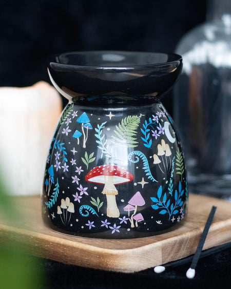 Close-up of the Dark Forest ceramic oil burner, with a black base adorned with colourful mushrooms, ferns, and stars, placed on a wooden surface with candles in the background.