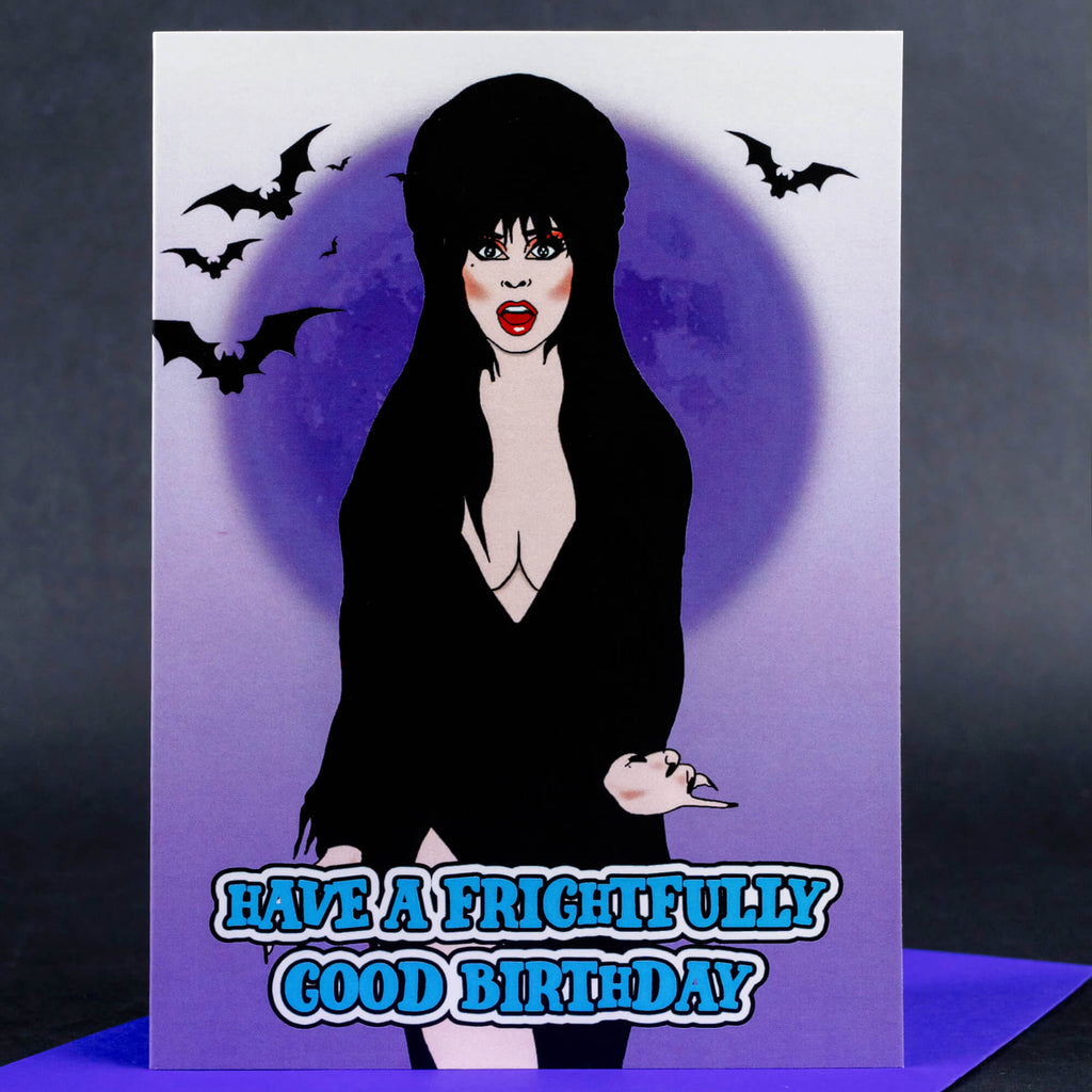 A spooky birthday card depicting a character inspired by Elvira with bats in the background and a glowing moon. The text reads “Have a frightfully good birthday” in bold, blue font, set against a purple gradient background.