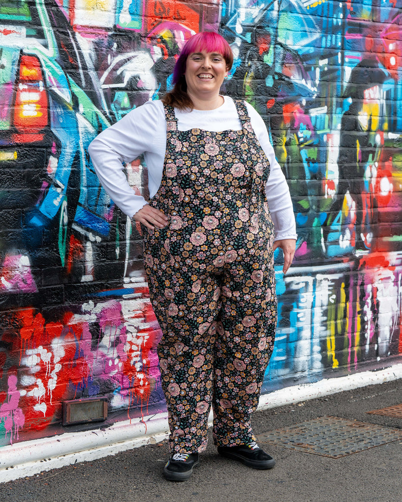 Full body shot of Chloe in the Eye Spy Floral Stretch Twill Dungarees, standing against graffiti, showing the alternative floral, skull, and eye design.