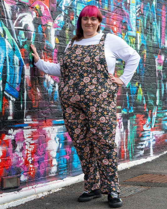 Chloe leans against a graffiti wall wearing Eye Spy Floral Stretch Twill Dungarees with a white long sleeve top, hands in pockets, smiling at the camera.