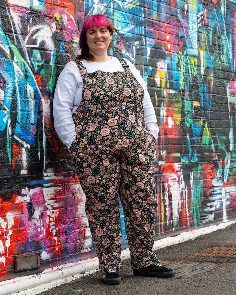 Chloe poses in the Eye Spy Floral Stretch Twill Dungarees with bold florals, skulls, and eyes, styled with a white top against colourful graffiti.