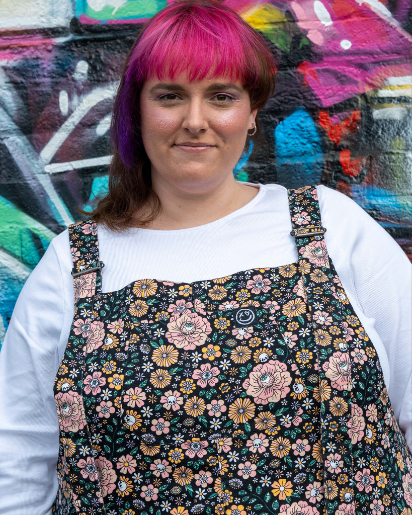 Chloe smiles in the Eye Spy Floral Stretch Twill Dungarees layered with a white top, showing the black twill covered in florals, eyes, and skulls.